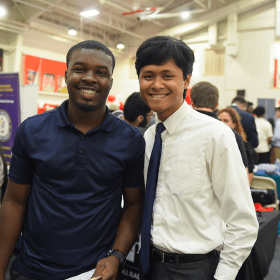 Two college students smiling at a campus career fair, standing side by side in a gymnasium with employer tables and attendees in the background. One student wears a navy polo shirt, and the other wears a white dress shirt with a tie, both appearing engaged and professional.