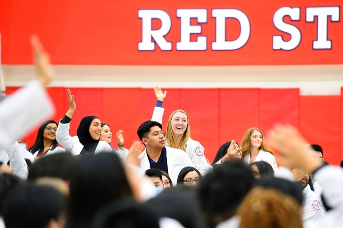 A group of St. John’s University Doctor of Pharmacy students wearing white lab coats raises their hands
